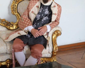 May include: A man wearing a black tricorn hat with a white feather, a pink and white patterned jacket, a gray vest, and brown pants sits on a gold and white ornate chair. He is holding his hands in his lap.