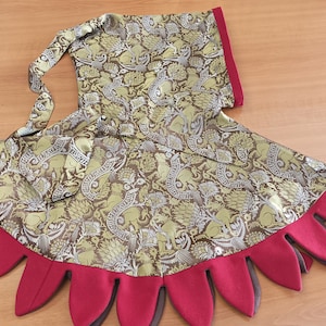 May include: A handmade costume piece featuring a patterned fabric in olive green, brown, and gold. The design includes a hood and a skirt-like bottom with red and brown petal-shaped accents. The fabric has a detailed floral and animal print.