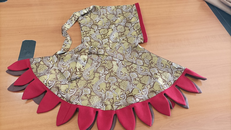 May include: A handmade costume piece featuring a patterned fabric in olive green, brown, and gold. The design includes a hood and a skirt-like bottom with red and brown petal-shaped accents. The fabric has a detailed floral and animal print.