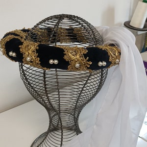 May include: A black velvet headband with gold trim and pearl accents. The headband is on a wire mannequin head with a white sheer fabric draped over it.