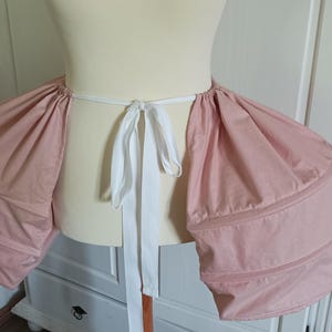 K&ouml;nnte beinhalten: Ein rosa-wei&szlig;er Stoff-Bustier mit einer wei&szlig;en Schleife. Das Bustier ist so konzipiert, dass es dem R&uuml;cken eines Rocks mehr Volumen verleiht.