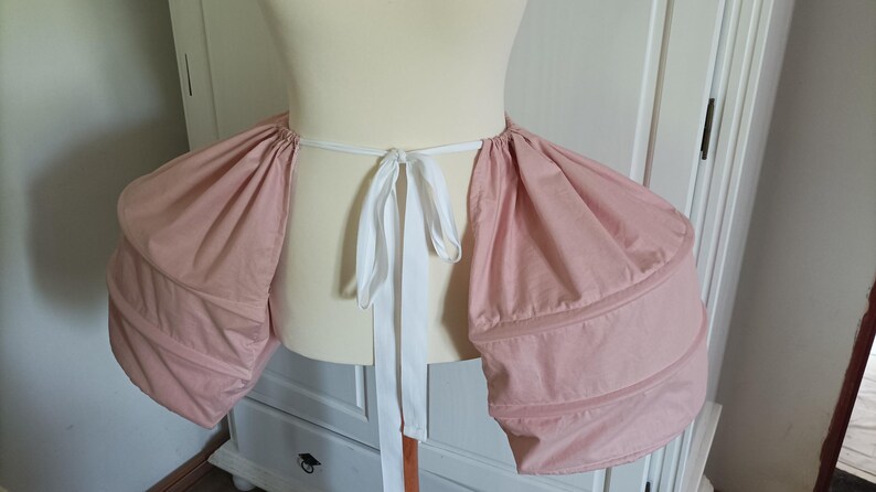 K&ouml;nnte beinhalten: Ein rosa-wei&szlig;er Stoff-Bustier mit einer wei&szlig;en Schleife. Das Bustier ist so konzipiert, dass es dem R&uuml;cken eines Rocks mehr Volumen verleiht.
