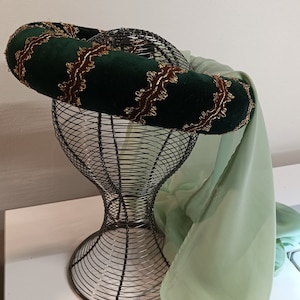 Puede incluir: Una diadema de terciopelo verde con bordados dorados, expuesta en una cabeza de maniquí de alambre. Una tela transparente verde claro a juego se drape sobre el maniquí.