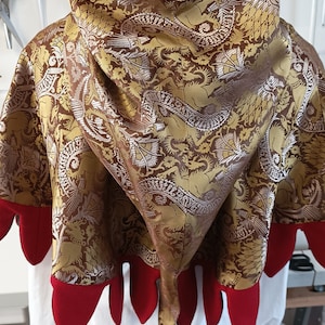 May include: A hooded cape with a dragon pattern in gold, brown, and silver. The hood and edges are trimmed with red fabric. The cape is displayed on a mannequin.
