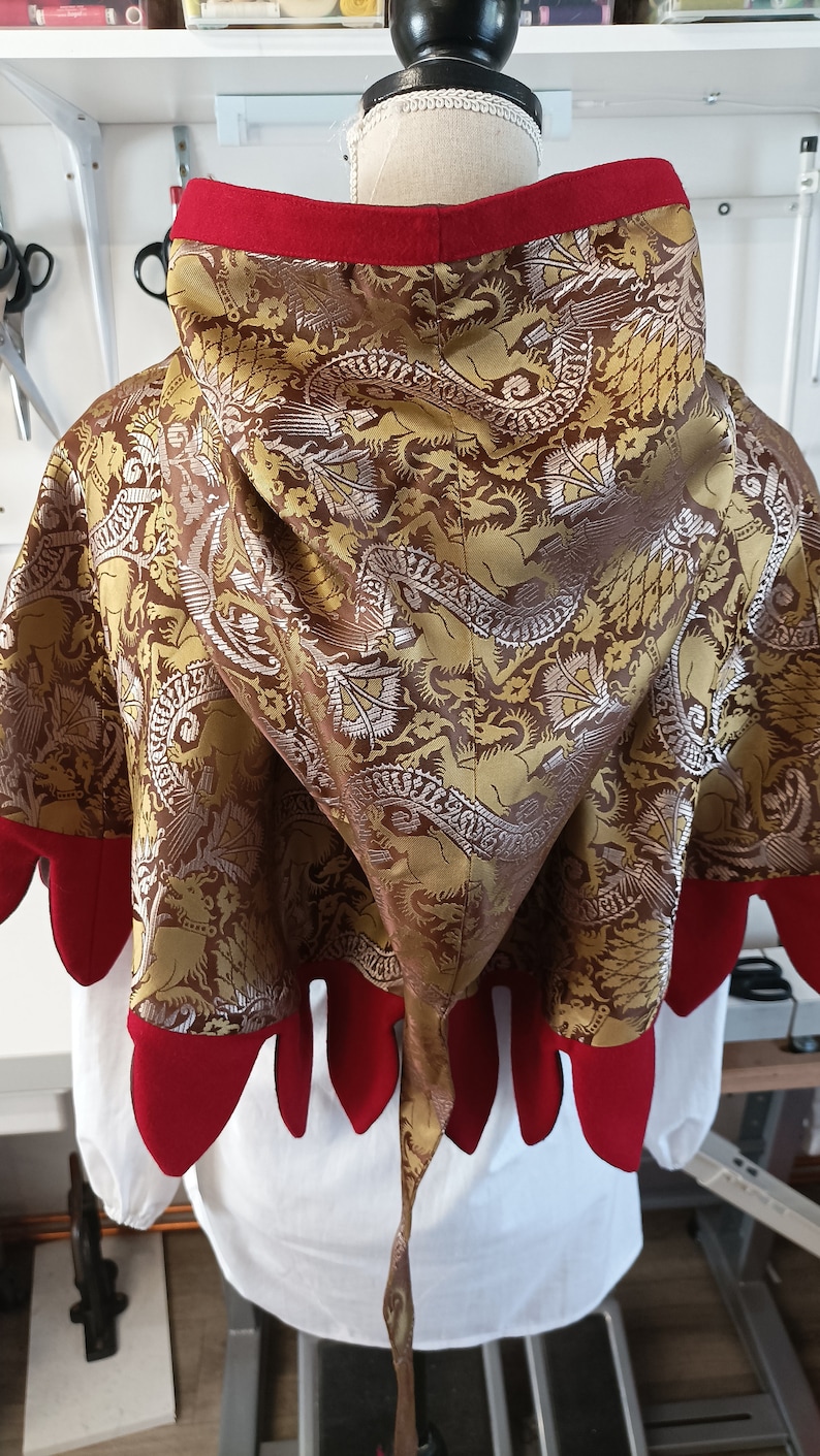 May include: A hooded cape with a dragon pattern in gold, brown, and silver. The hood and edges are trimmed with red fabric. The cape is displayed on a mannequin.