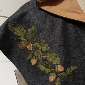 Op de afbeelding: Een grijze capuchonmantel met een bruine voering en een geborduurd ontwerp van groene eikenbladeren en bruine eikels.