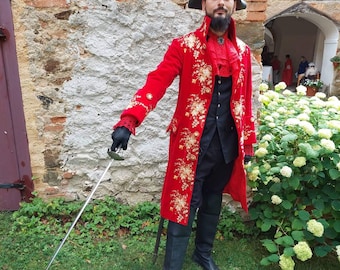 May include: A man in a red embroidered coat with gold accents, a black vest, and black pants. He is wearing a black hat and holding a sword. He is standing in front of a brick wall with green grass and white flowers in the background.