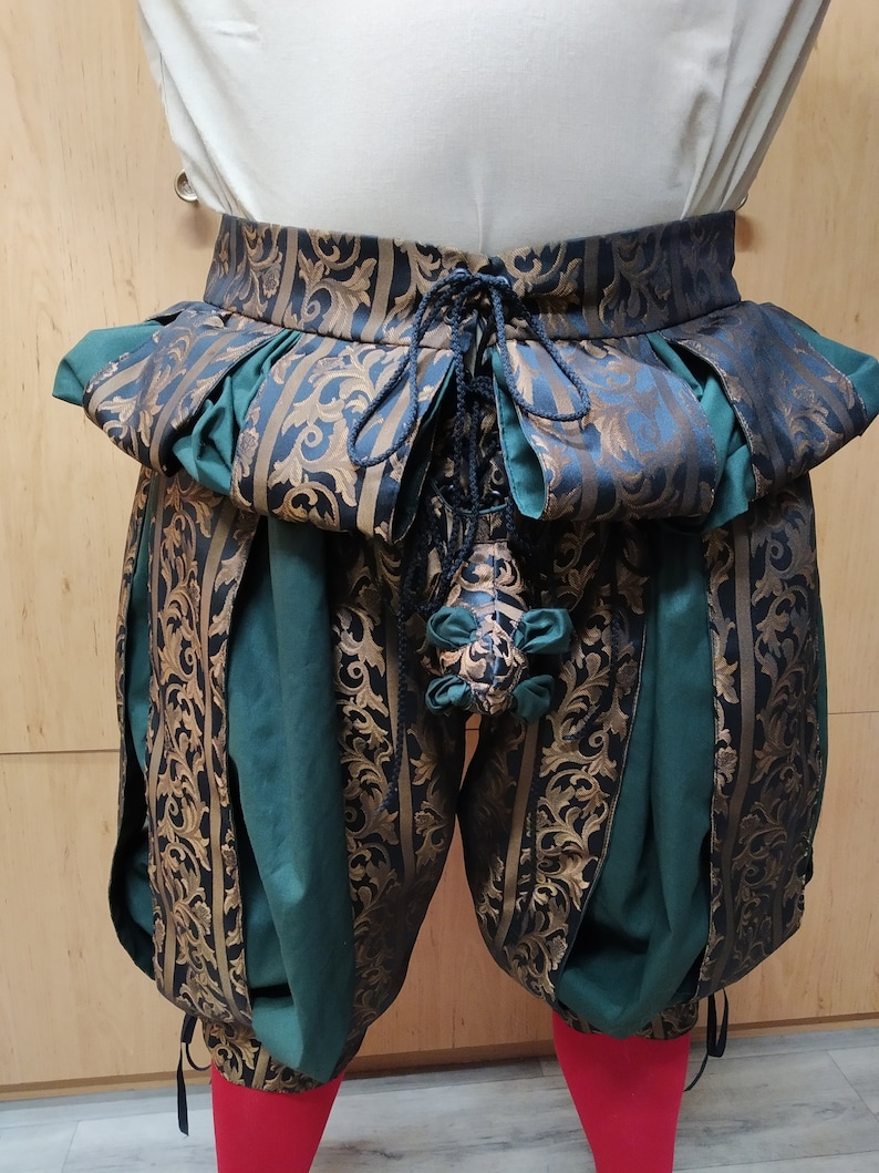 May include: Close-up of Renaissance-style breeches. The breeches feature a high waist with a pleated, patterned band in teal and gold. The main body is green with gold floral brocade panels. A drawstring and decorative tassel are visible.