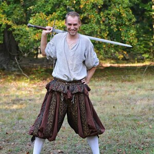 May include: A person wearing a light gray tunic and brown and black patterned breeches, holding a long sword. The outfit is reminiscent of historical attire. The person is standing in a grassy area with trees in the background.