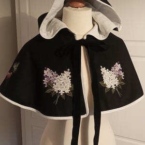 Può includere: Un mantello nero con cappuccio, bordatura bianca e un nastro di velluto nero. Il mantello presenta fiori di lillà ricamati in bianco e viola.