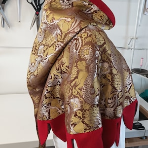 May include: A decorative hooded cloak with a brown and gold patterned fabric, featuring a red trim and pointed edges. The cloak is displayed on a mannequin, showcasing its design and construction. The background includes sewing tools.