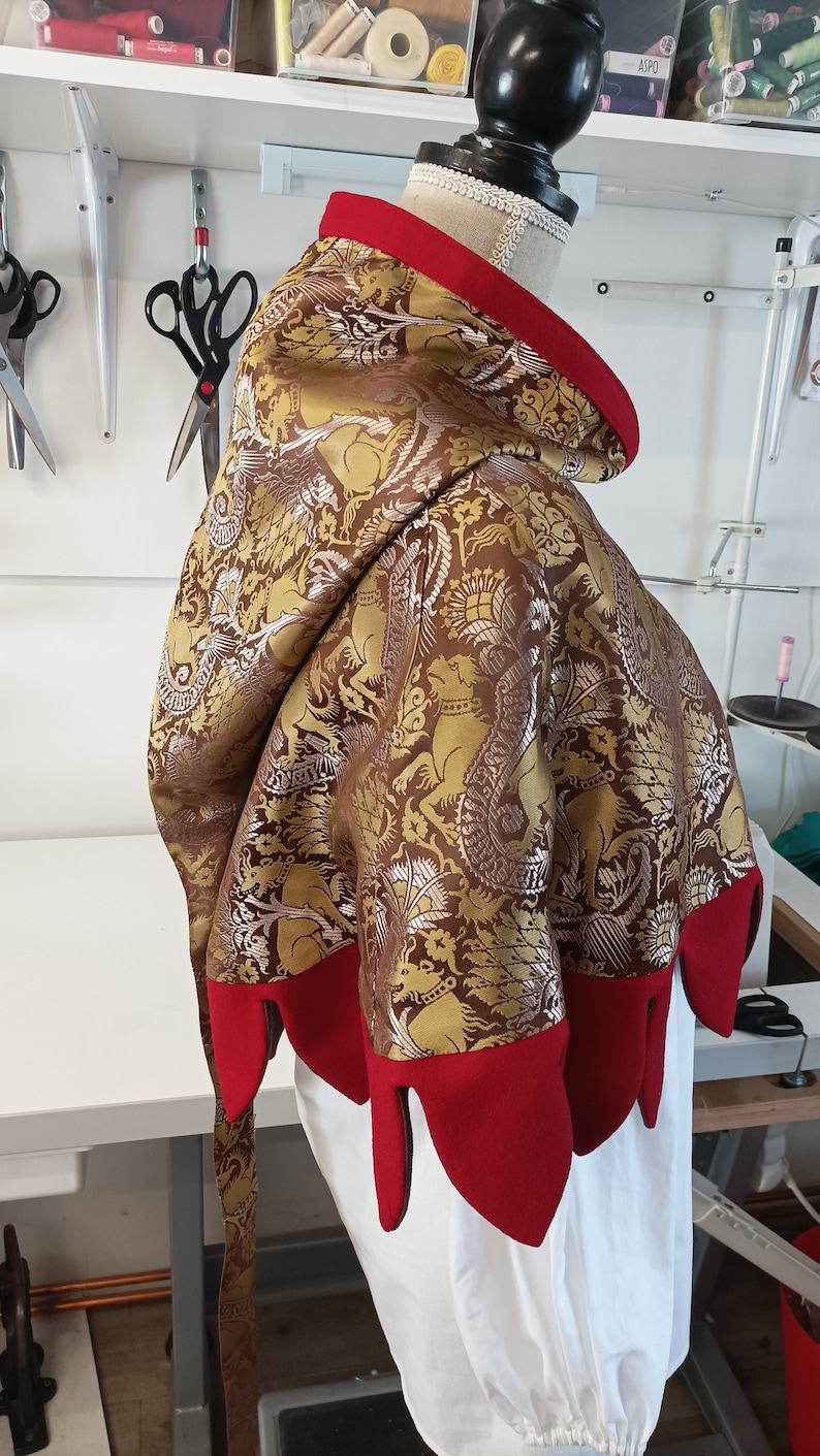 May include: A decorative hooded cloak with a brown and gold patterned fabric, featuring a red trim and pointed edges. The cloak is displayed on a mannequin, showcasing its design and construction. The background includes sewing tools.