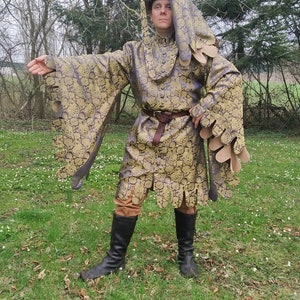 May include: A person wearing a gold and purple patterned robe with a large hood. The robe has long, flowing sleeves with decorative fringe. The person is standing in a grassy field.