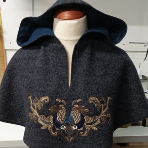May include: A dark gray hooded cape with a deep V-neckline. The cape features an embroidered design of two stylized birds in gold, blue, and brown. The hood is lined with a contrasting blue fabric.