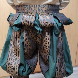 May include: A Renaissance-style costume featuring green and gold patterned breeches with a matching belt. The breeches have a puffed design and are paired with a cream-colored shirt. The belt and breeches have a floral pattern.