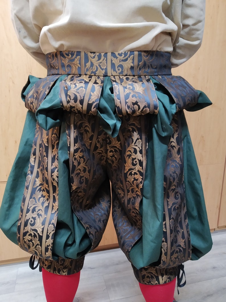 May include: A Renaissance-style costume featuring green and gold patterned breeches with a matching belt. The breeches have a puffed design and are paired with a cream-colored shirt. The belt and breeches have a floral pattern.