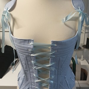 May include: A light blue corset with a lace-up back and ribbon detail. The corset is made of quilted fabric and has a fitted silhouette.