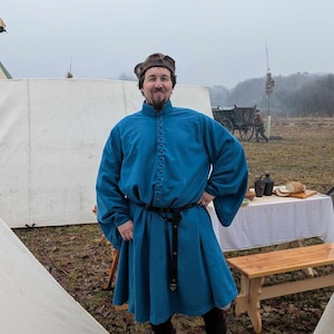 May include: A person wearing a teal medieval tunic with long sleeves and a button-down front. The tunic is paired with a black belt and boots. A brown hat is worn. The background includes a white tent, a wooden table, and a cart.