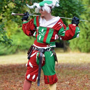 Könnte beinhalten: Eine Person in einem grün-roten Kostüm mit einem weißen Federhut hält ein Schwert vor sich. Die Person steht in einem grasbewachsenen Bereich mit heruntergefallenen Blättern.