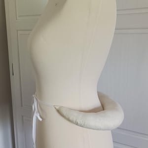 Könnte beinhalten: Eine cremefarbene Schneiderpuppe mit gepolstertem Hüftpolster. Die Puppe steht vor einem weißen Hintergrund mit einer weißen Tür und einem Schrank. Ein weißes Band ist um die Taille der Puppe gebunden.