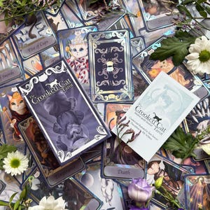 May include: A deck of The Crooked Cat Tarot cards featuring purple and silver holographic accents, a matching box with a cat illustration, and an intuitive guide booklet, all surrounded by white daisies, purple roses, and green foliage.