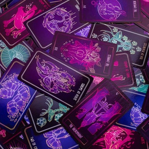 May include: A collection of tarot cards with a dark, mystical theme. Each card features intricate illustrations in shades of purple, pink, and teal, with gold accents. The cards display titles such as "The Emperor," "The Empress," and "Seven of Swords."