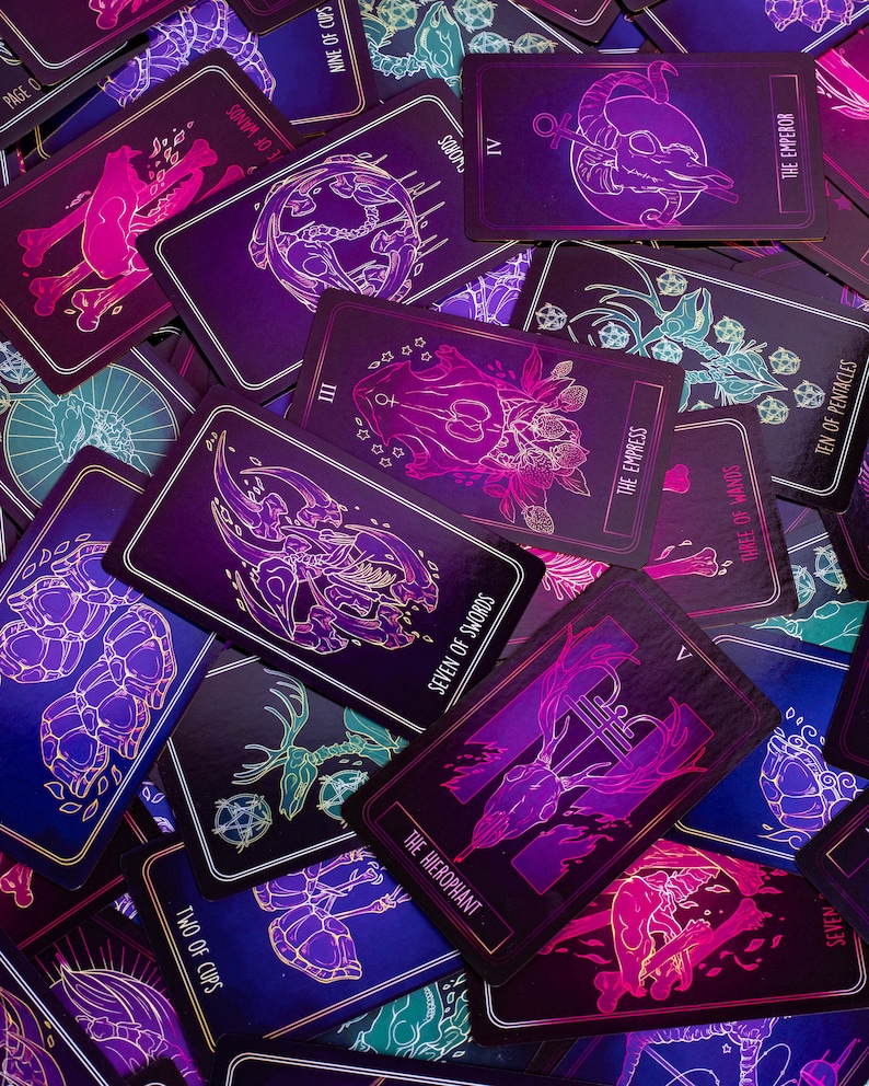 May include: A collection of tarot cards with a dark, mystical theme. Each card features intricate illustrations in shades of purple, pink, and teal, with gold accents. The cards display titles such as "The Emperor," "The Empress," and "Seven of Swords."