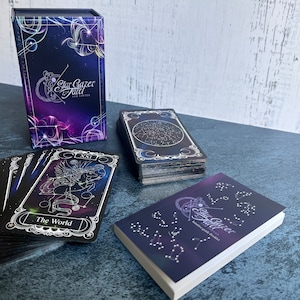 May include: A Stargazer Tarot deck featuring a purple and blue box with celestial graphics, a stack of cards with a star map design, a single card labeled The World with a winged horse illustration, and a small booklet with constellation patterns.