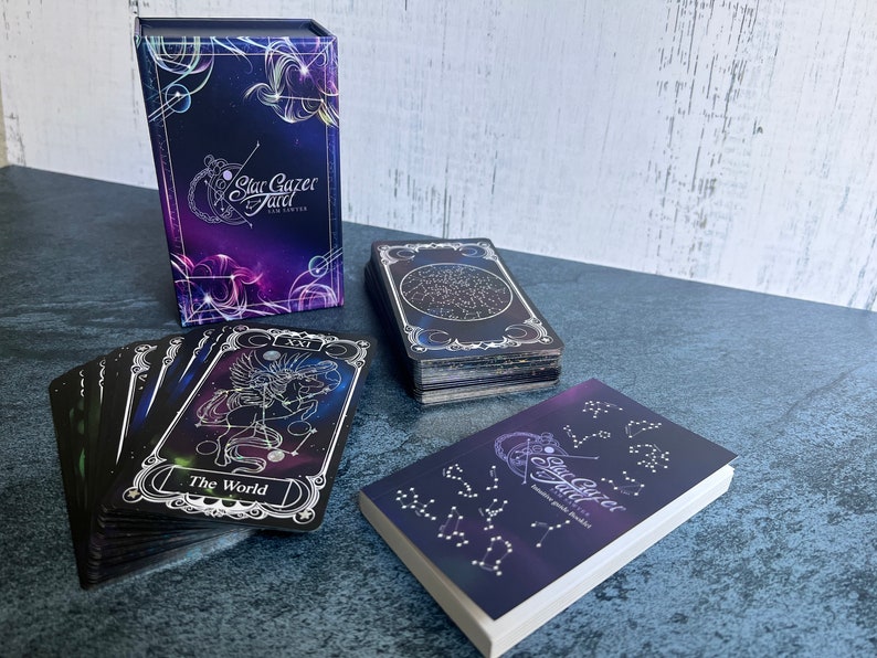 May include: A Stargazer Tarot deck featuring a purple and blue box with celestial graphics, a stack of cards with a star map design, a single card labeled The World with a winged horse illustration, and a small booklet with constellation patterns.