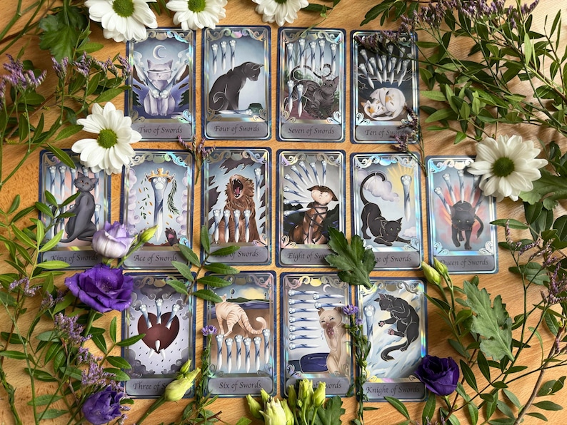 May include: A collection of fourteen tarot cards from a cat-themed deck, each featuring unique feline illustrations and sword motifs. The cards are arranged in a grid, displaying titles like Two of Swords, Four of Swords, and Queen of Swords.