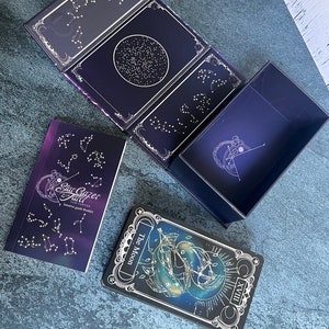 May include: A Stargazer Tarot deck set featuring a dark purple box with gold constellation illustrations, an intuitive guide booklet with the text Stargazer Tarot Sam Sawyer, and a tarot card titled The Moon with the Roman numeral XVIII.