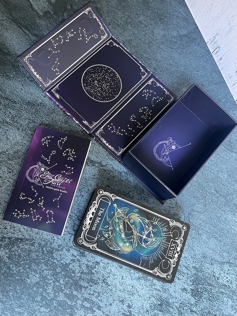 May include: A Stargazer Tarot deck set featuring a dark purple box with gold constellation illustrations, an intuitive guide booklet with the text Stargazer Tarot Sam Sawyer, and a tarot card titled The Moon with the Roman numeral XVIII.