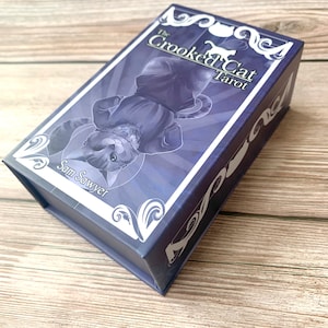 May include: A rectangular purple tarot card deck box featuring an illustration of a cat lying on its back. The text on the box reads The Crooked Cat Tarot and Sam Sawyer. The box has white decorative scrollwork patterns on the corners and edges.