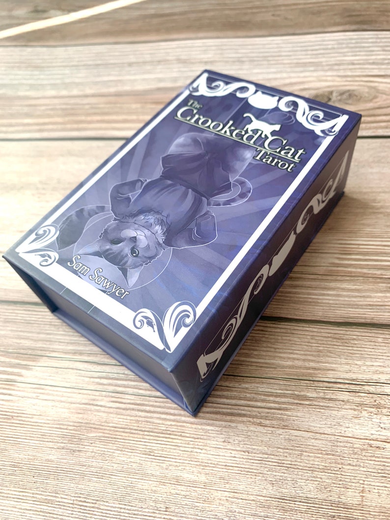 May include: A rectangular purple tarot card deck box featuring an illustration of a cat lying on its back. The text on the box reads The Crooked Cat Tarot and Sam Sawyer. The box has white decorative scrollwork patterns on the corners and edges.