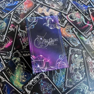 May include: A rectangular deck box for the Stargazer Tarot by Sam Sawyer, featuring a deep purple and blue nebula background with white celestial line art. The box is surrounded by multiple tarot cards with similar cosmic designs.