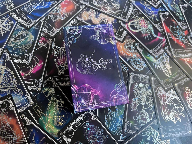 May include: A rectangular deck box for the Stargazer Tarot by Sam Sawyer, featuring a deep purple and blue nebula background with white celestial line art. The box is surrounded by multiple tarot cards with similar cosmic designs.