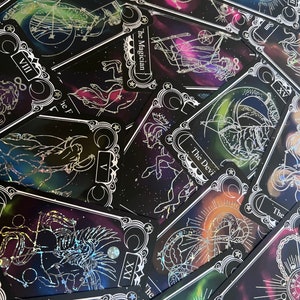 May include: A collection of tarot cards featuring black backgrounds with colorful nebula-like patterns and silver foil line art illustrations. Visible cards include The Magician, The Devil, The Empress, The Moon, The Sun, and Wheel of Fortune.