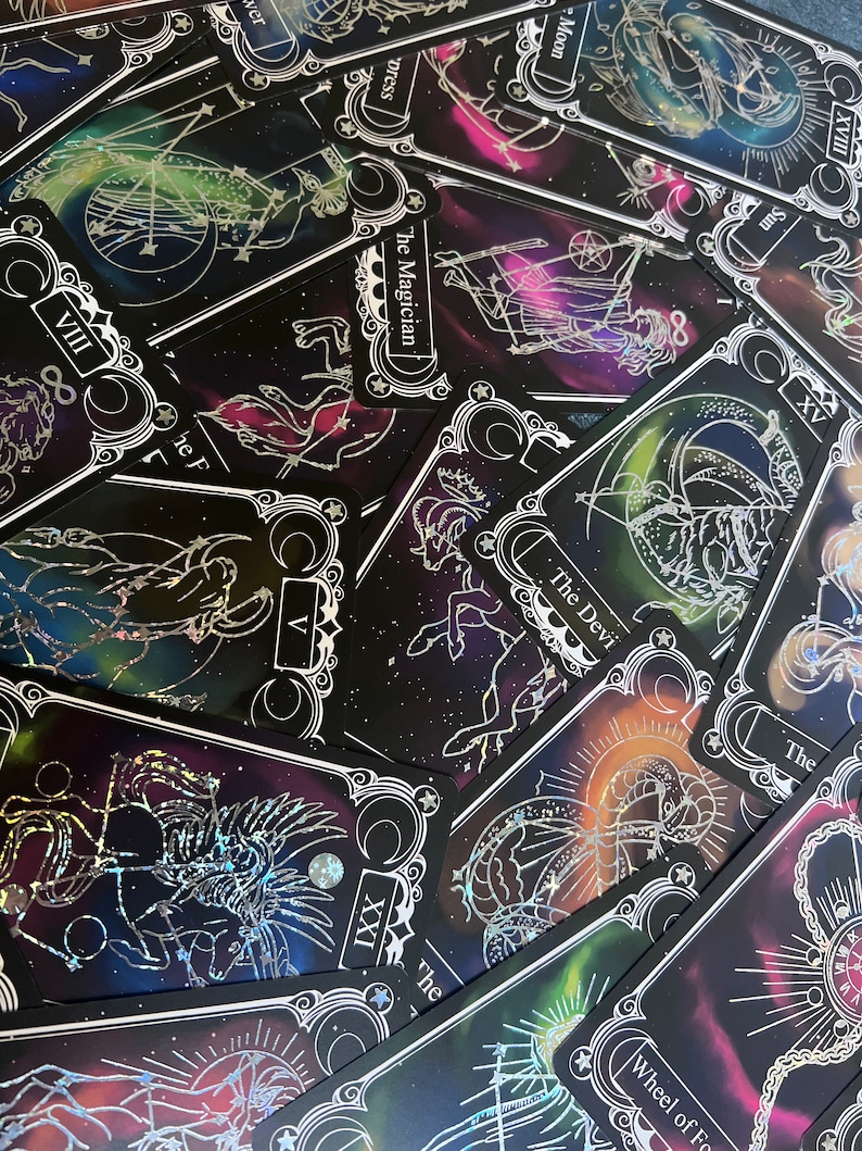 May include: A collection of tarot cards featuring black backgrounds with colorful nebula-like patterns and silver foil line art illustrations. Visible cards include The Magician, The Devil, The Empress, The Moon, The Sun, and Wheel of Fortune.