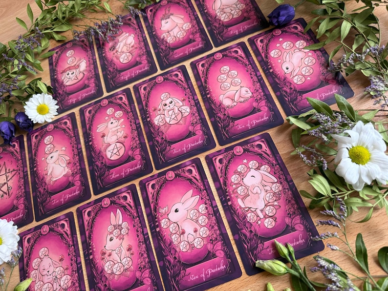 May include: A collection of tarot cards featuring illustrations of rabbits and pentacles in shades of pink and purple. Each card displays a unique rabbit character surrounded by floral motifs and pentacle symbols, with text labels at the bottom.