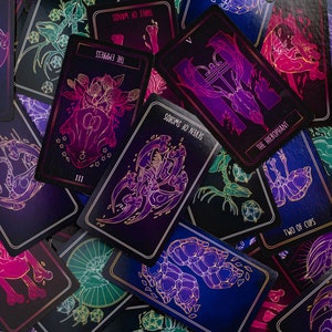 May include: A collection of tarot cards with dark backgrounds and neon illustrations. The cards feature various symbols and text, including "The Empress," "Seven of Swords," and "Two of Cups." The color palette includes purple, pink, blue, and green.