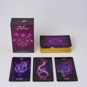 May include: A tarot card deck titled "The Bone Tarot" with a purple and gold color scheme. The deck includes a box, a deck of cards with gold edges, and three individual cards. The box and cards feature illustrations and the text "The Bone Tarot".