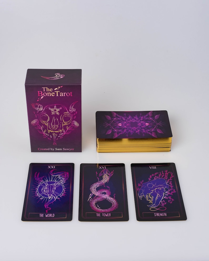 May include: A tarot card deck titled "The Bone Tarot" with a purple and gold color scheme. The deck includes a box, a deck of cards with gold edges, and three individual cards. The box and cards feature illustrations and the text "The Bone Tarot".
