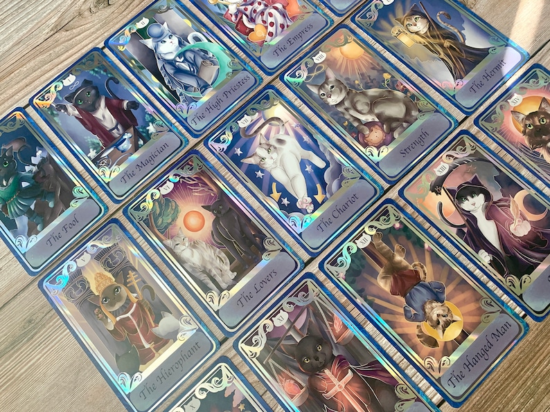 May include: A collection of rectangular tarot cards featuring cat illustrations with holographic borders. Each card displays a feline character in a specific role, including The Magician, The High Priestess, The Empress, The Hermit, Strength, The Chariot, The Fool, The Lovers, The Hierophant, The Hanged Man, and Death.