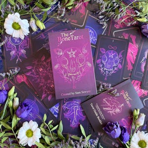 Peut inclure: Un jeu de cartes de tarot et un livret intitulé 'The Bone Tarot', avec un design violet et noir. Les cartes et le livre présentent des illustrations de crânes d'animaux et de motifs floraux. L'image est ornée de fleurs et de feuillage.