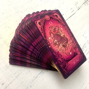 May include: A deck of tarot cards fanned out, revealing the card "The Fool". The card features a purple background with a whimsical illustration of a fox wearing a jester's hat and surrounded by stars and ribbons.