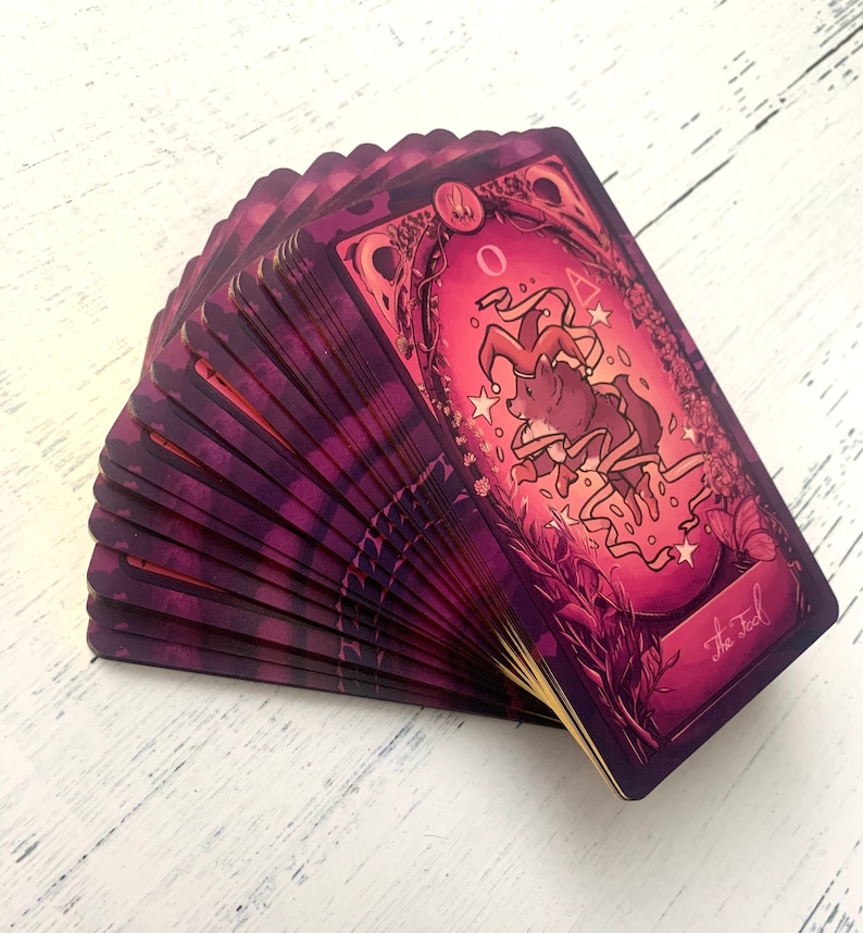 May include: A deck of tarot cards fanned out, revealing the card "The Fool". The card features a purple background with a whimsical illustration of a fox wearing a jester's hat and surrounded by stars and ribbons.