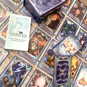May include: A deck of The Crooked Cat Tarot cards featuring feline-themed illustrations, a purple storage box with cat artwork, and an intuitive guide booklet. The cards display various scenes with cats, including The Tower, The Devil, and The Lovers.