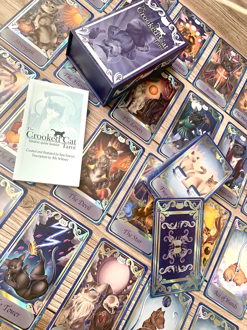 May include: A deck of The Crooked Cat Tarot cards featuring feline-themed illustrations, a purple storage box with cat artwork, and an intuitive guide booklet. The cards display various scenes with cats, including The Tower, The Devil, and The Lovers.