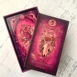 May include: A closed box of The Fae and the Ferns Tarot cards by Samantha Sawyer. The box is purple with a skull and floral design. The text on the box reads "The Fae and the Ferns Tarot" and "by Samantha Sawyer Sincerely Sam".