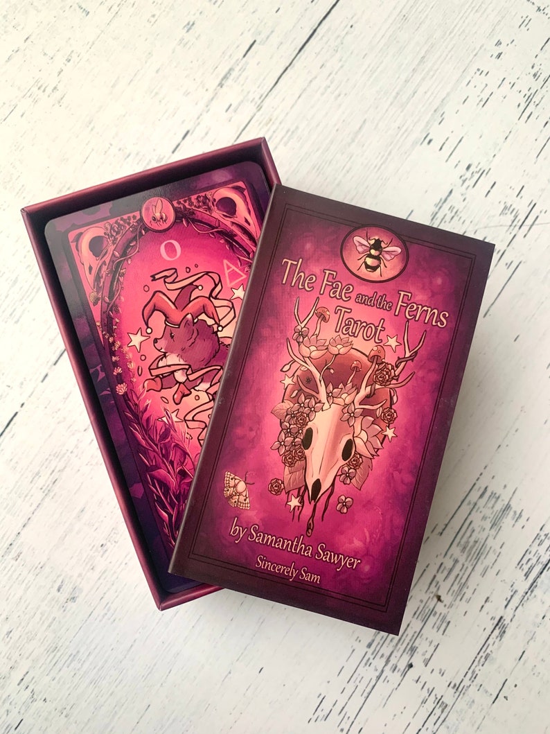 May include: A closed box of The Fae and the Ferns Tarot cards by Samantha Sawyer. The box is purple with a skull and floral design. The text on the box reads "The Fae and the Ferns Tarot" and "by Samantha Sawyer Sincerely Sam".